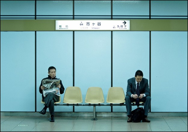 one man reads a newspaper; another reads his phone